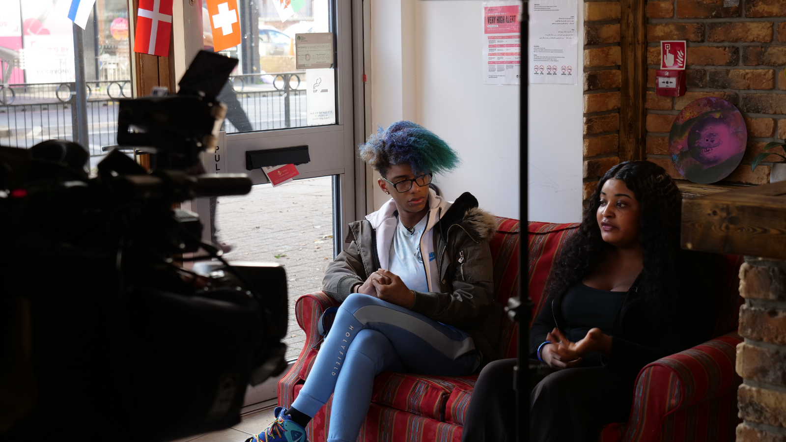 Two young people sit on a sofa while talking to a camera filming them.